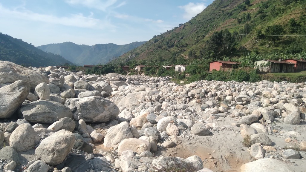 A river bed in Kasese filled with boulders and debris after flooding, with homes and the Rwenzori slopes behind